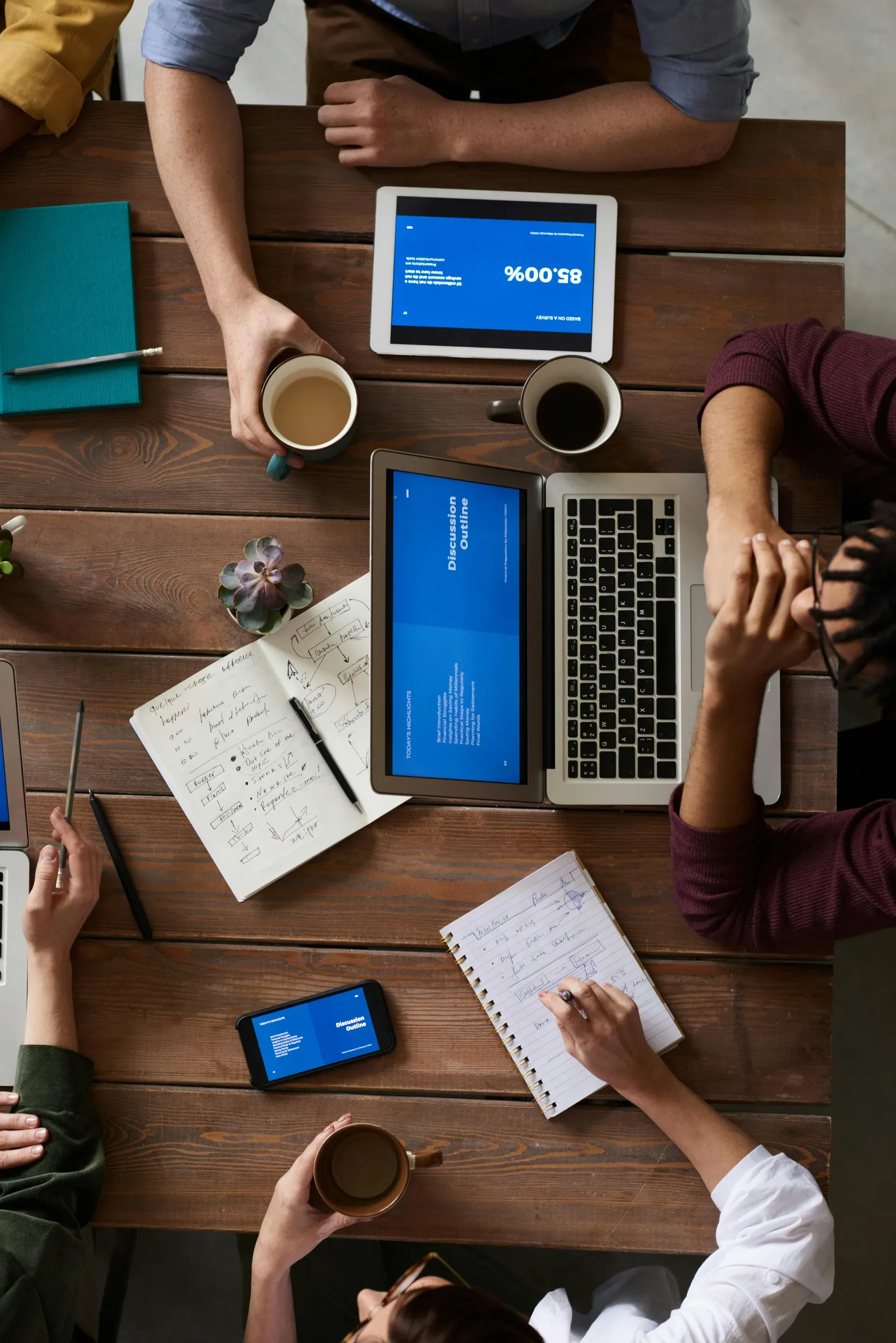 Overhead view of four colleagues around a wooden table, with laptops and a tablet showing presentation slides, notebooks and coffee cups