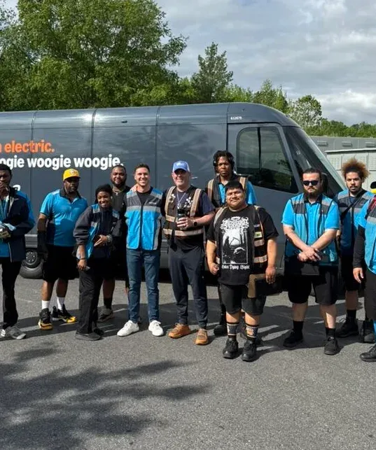 A team of delivery drivers in blue vests gathered in front of an electric delivery van