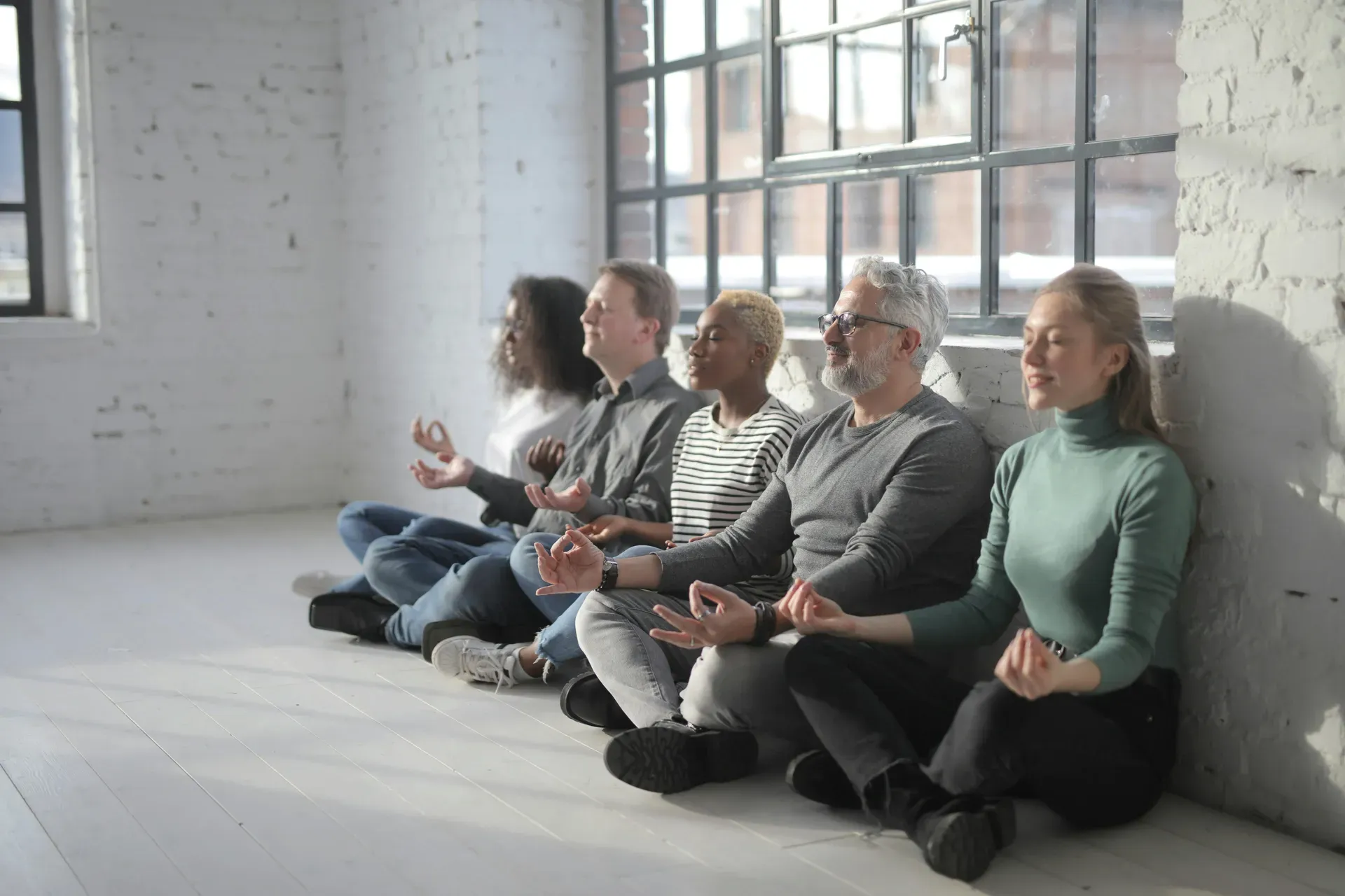 Five colleagues sit cross-legged along a white brick wall with their eyes closed and hands resting on their knees