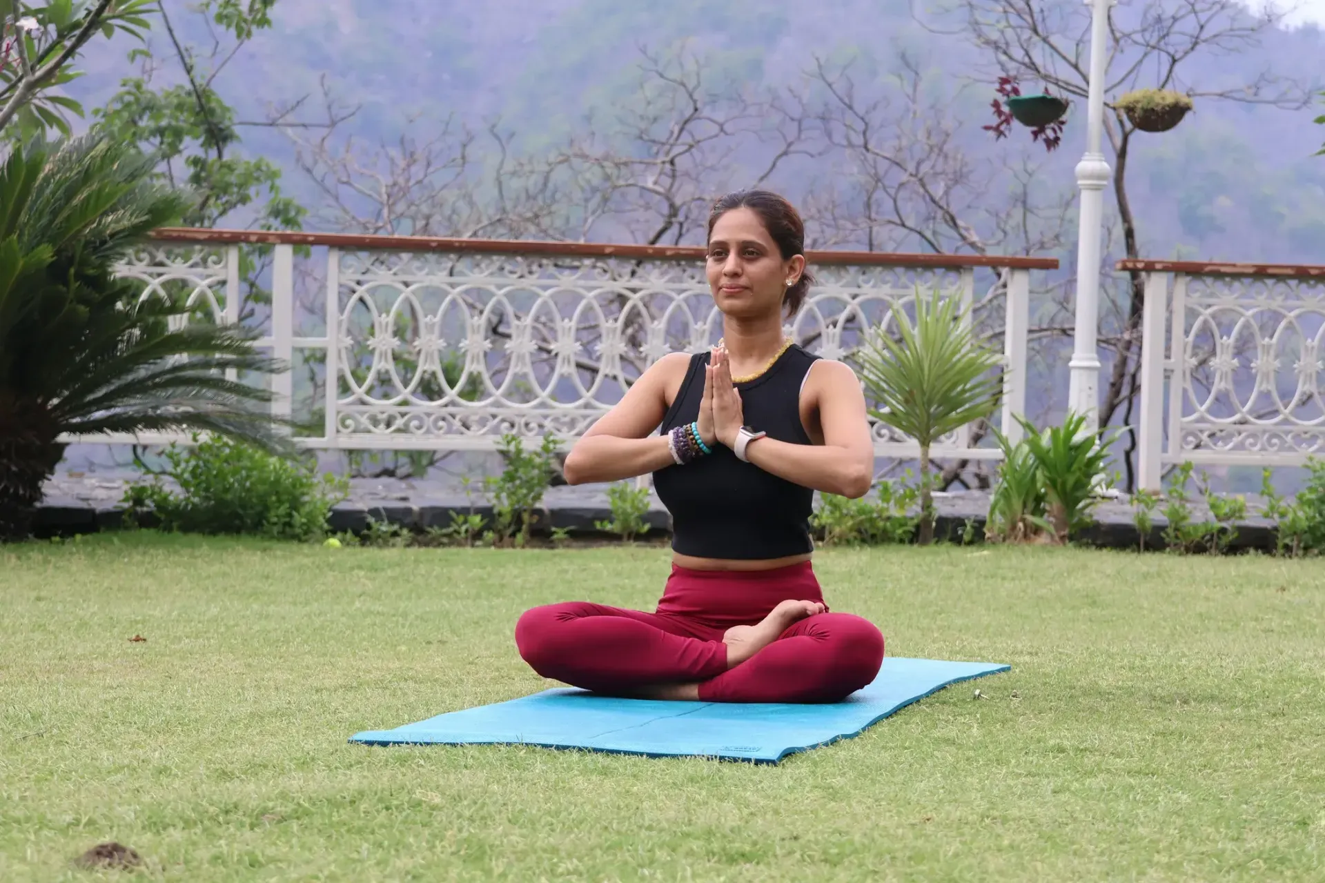 A woman sits cross-legged on a mat on a lawn, palms pressed together, with hills behind her