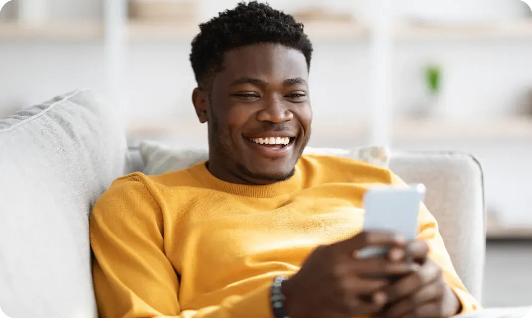 Smiling young man in a yellow sweater sitting on a couch using a smartphone.