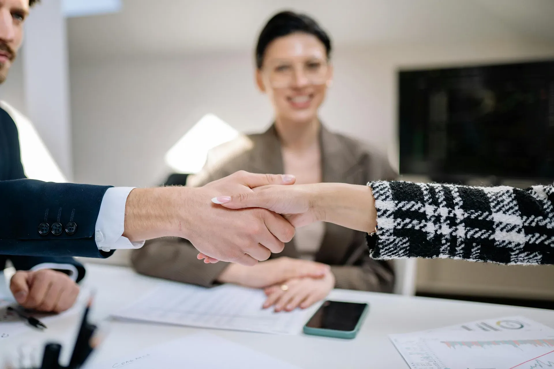 Two people shake hands across a desk while a third looks on, with printed charts on the table