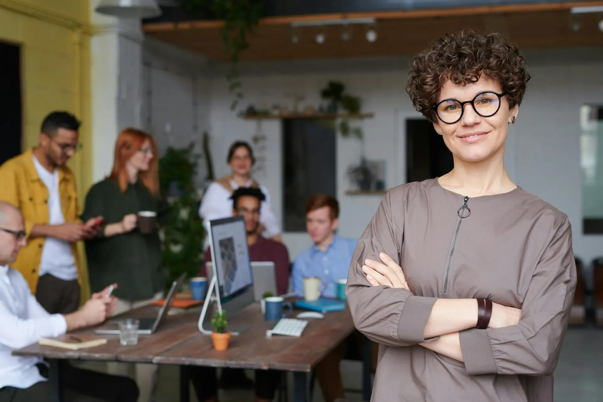 A woman in glasses stands with her arms folded, smiling, while colleagues work at a table behind her