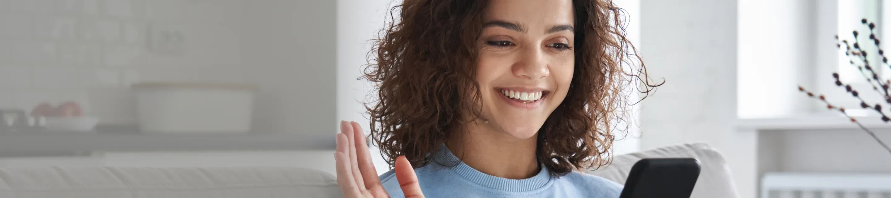 Smiling woman with curly hair waving while looking at a smartphone in a bright living room.