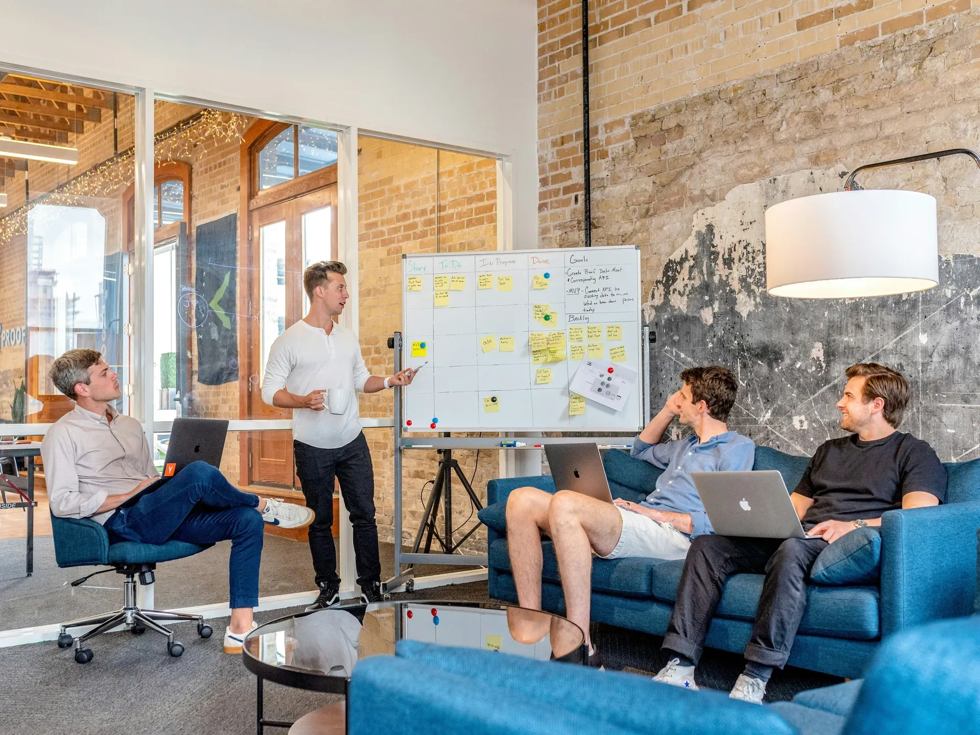 A man stands beside a whiteboard covered in sticky notes, talking to three colleagues seated on sofas