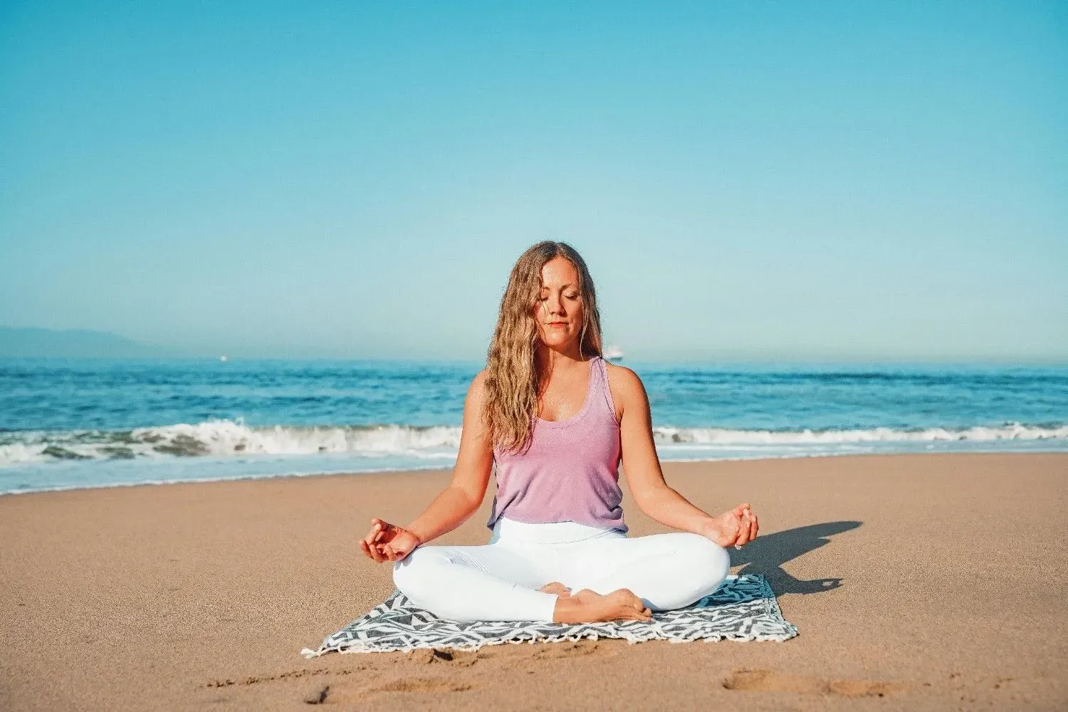 A woman sits cross-legged on a towel on the sand, eyes closed, with the ocean behind her