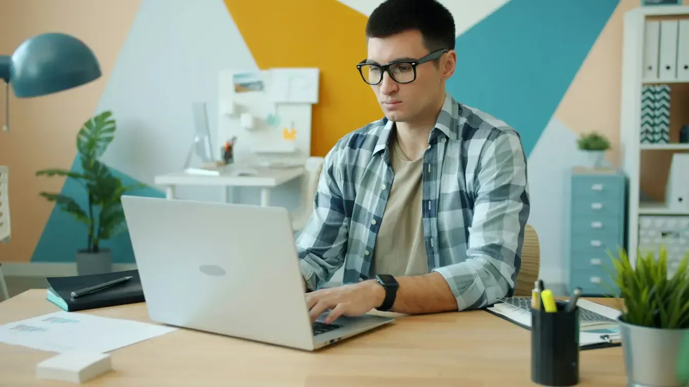 A man in glasses works at a laptop at a tidy desk, in front of a wall painted in overlapping colour blocks