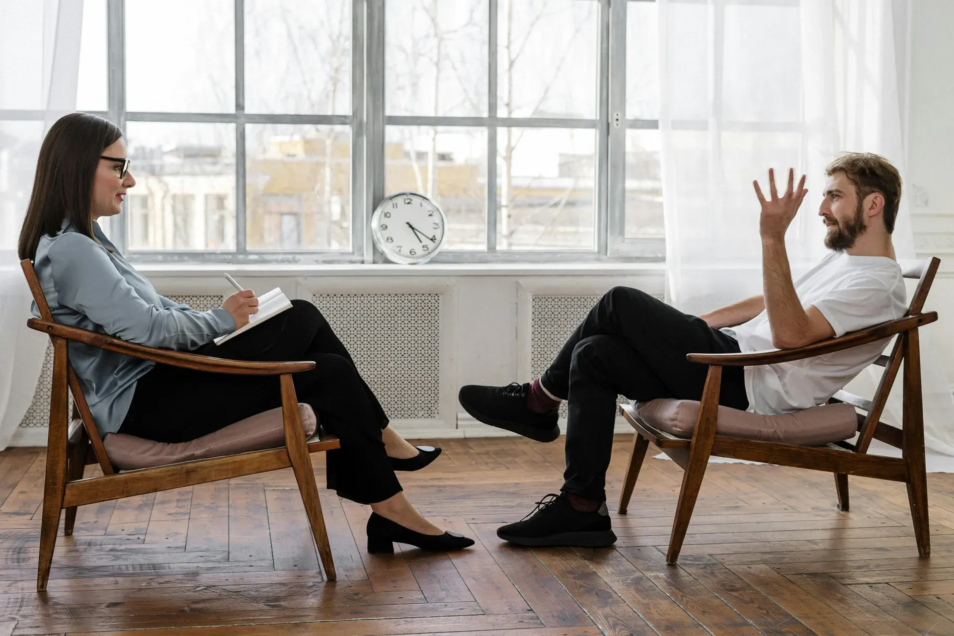 Two people sit facing each other in wooden chairs by a bright window, one taking notes while the other talks