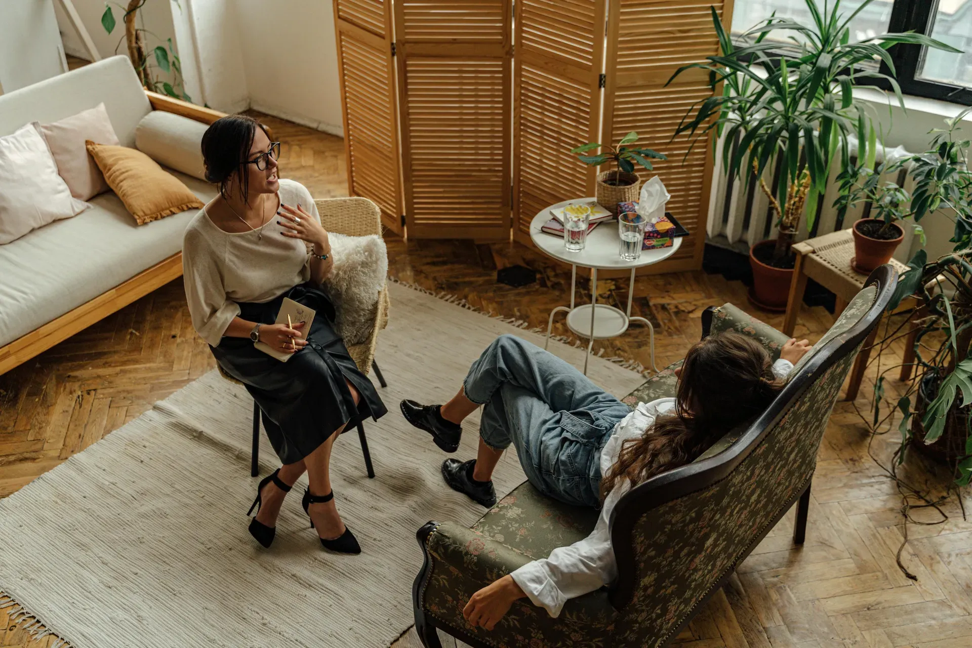 Overhead view of a woman with a notebook talking to another person reclining on a chaise in a plant-filled room