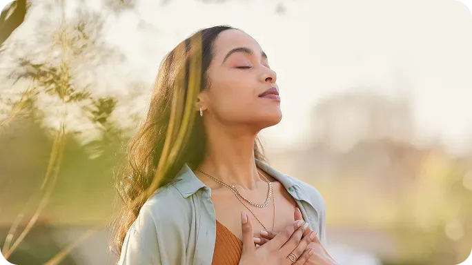 Woman with eyes closed, hands over her chest, standing peacefully outdoors with sunlight in the background.
