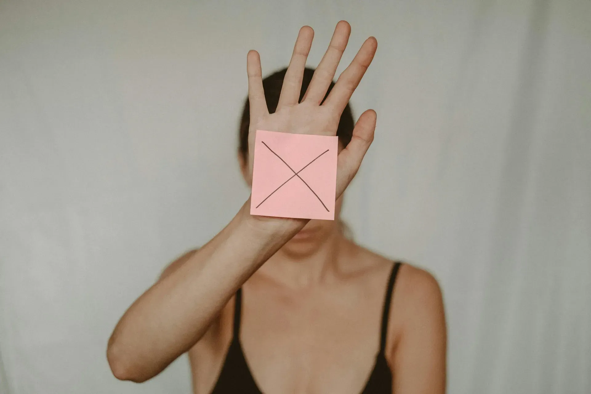 A person holds a palm towards the camera, a sticky note marked with a cross covering their face