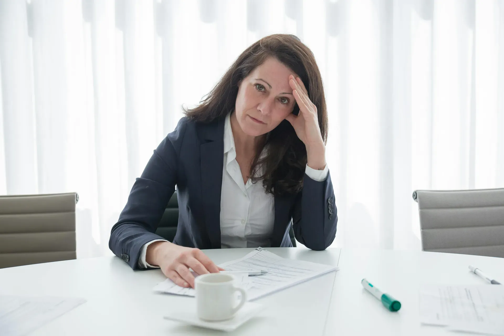 A woman in a blazer sits at a white table resting her head against her hand, papers and a coffee cup in front of her