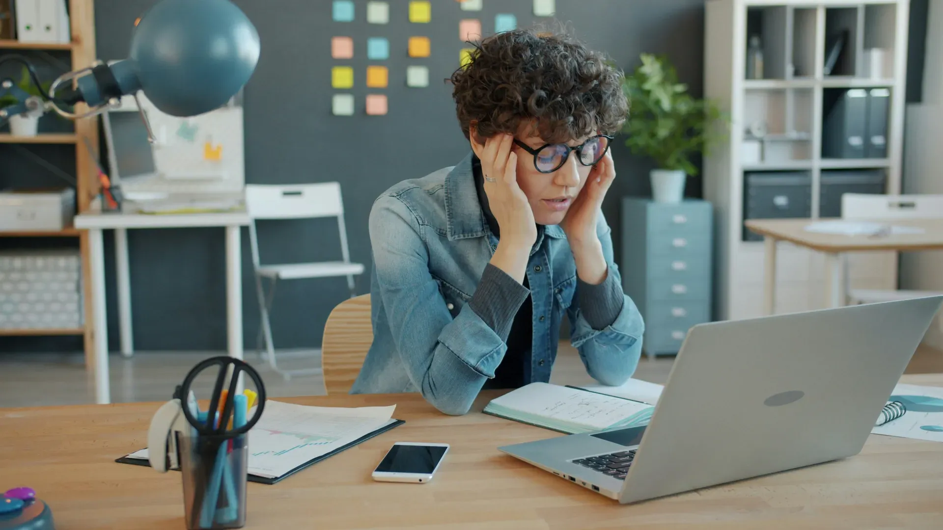 A woman at a desk holds her head in both hands as she looks at her laptop, sticky notes on the wall behind her