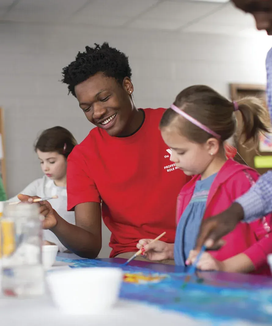 A YMCA staff member in a branded red shirt painting at a table with a group of children