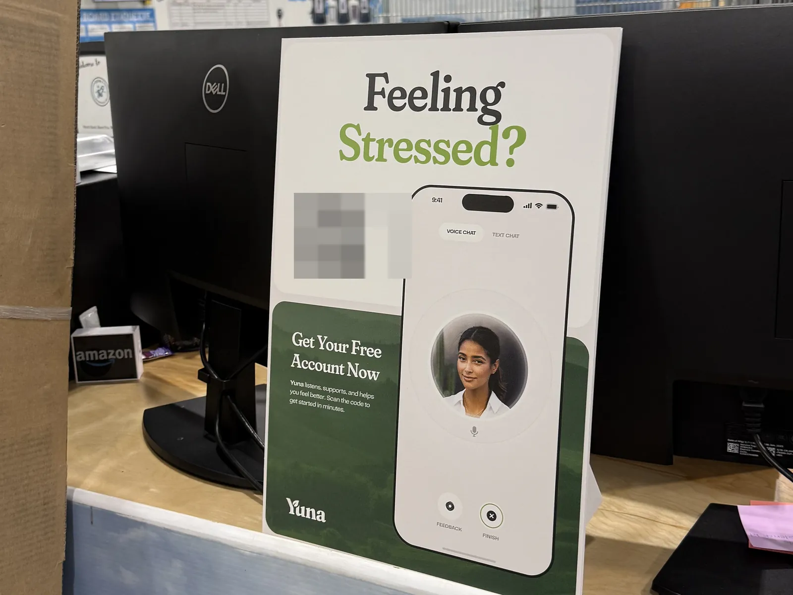 A Yuna poster standing on a desk between monitors inside a delivery station, reading Feeling Stressed? with a phone showing the app